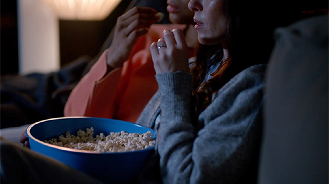 Two people sitting on a couch with a bowl of popcorn and a piece of popcorn in hand ready to eat